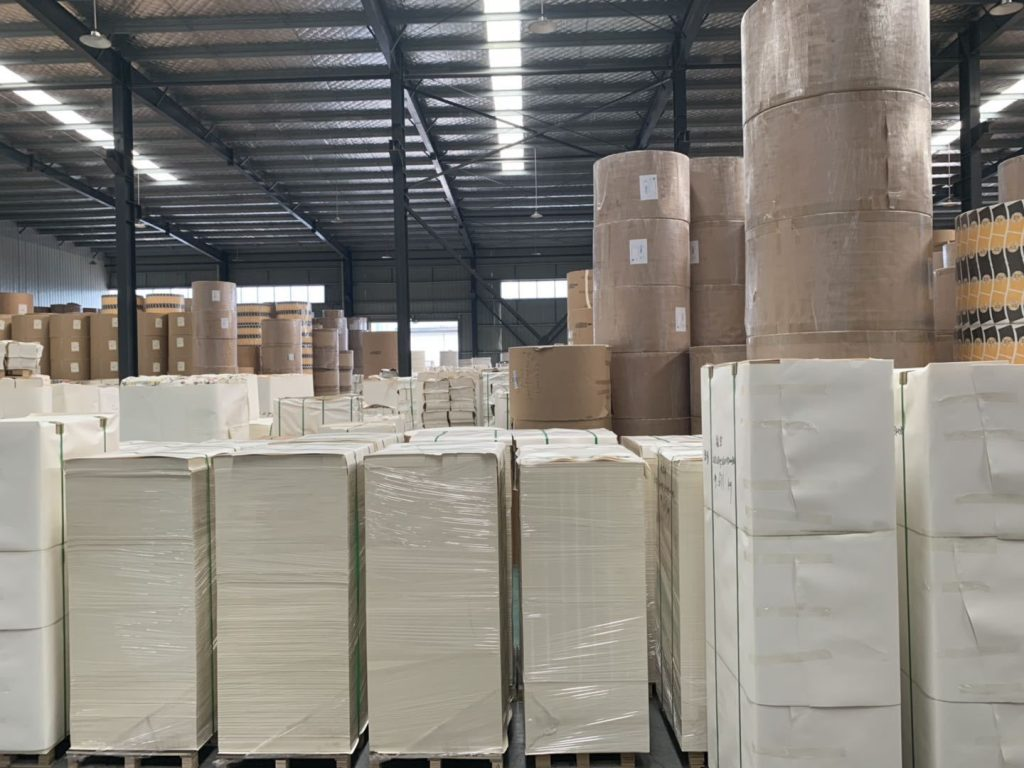 A large paper packaging warehouse storing massive kraft paper rolls and neatly stacked paperboard sheets for custom eco friendly food packaging manufacturing.
