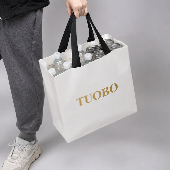 A person carrying a sturdy white paper bag with black handles and a gold logo, loaded with beverage cans and plastic water bottles.