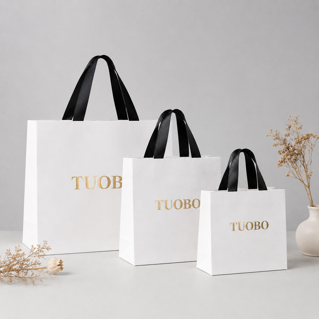 Three white paper gift bags in small, medium, and large sizes featuring gold foil logos and black ribbon handles on a beige table.