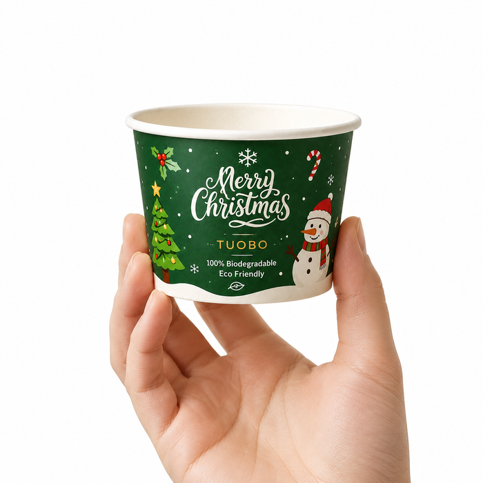Hand holding size reference shot of TUOBO green Christmas biodegradable paper ice cream cup, decorated with snowman, Christmas tree, candy cane holiday graphics, eco friendly compostable disposable dessert container for wholesale seasonal food packaging