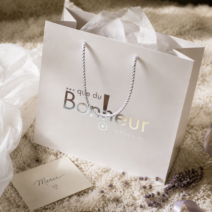 A white gift bag with rope handles and metallic foil logo on a cozy rug next to a thank you card.