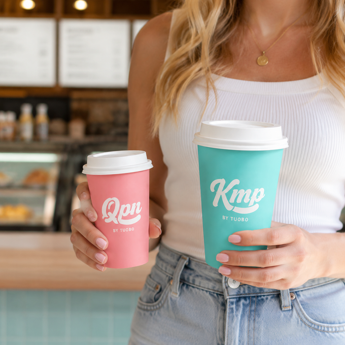 A woman holding two custom takeaway coffee cups in different sizes with white lids inside a cafe.