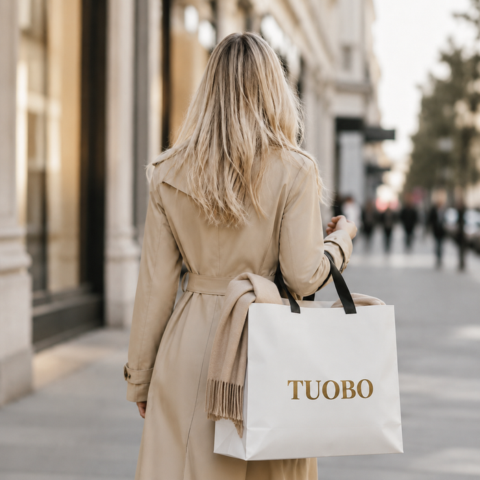 Rear view of a woman in a beige trench coat walking outdoors while holding a white luxury paper shopping bag with black handles and a gold TUOBO logo.