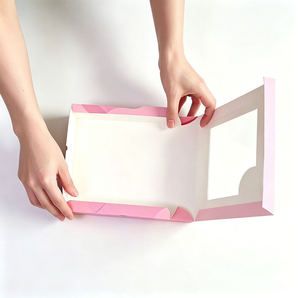 Top-down shot of hands unfolding TUOBO matte pink paper cupcake box to show folding structure, blank interior, clear PVC window design, disposable customizable dessert packaging for wholesale