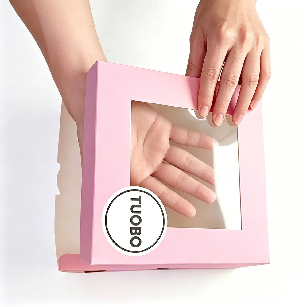 White background product shot of two hands holding TUOBO matte pink paper cupcake box with clear viewing window, custom logo printable bakery takeaway box for small desserts wholesale