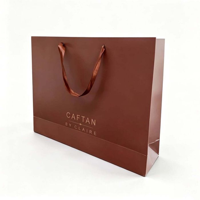 Side view of a brown custom paper bag featuring satin ribbon handles and a stamped rose gold logo.