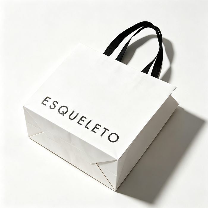 High-angle view of a white designer paper shopping bag with black handles resting on a light surface.