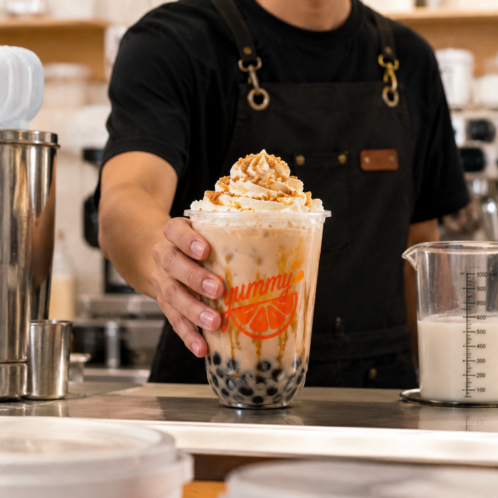 Barista handing creamy bubble tea in custom branded clear rPET U shaped cups, food grade transparent recycled plastic disposable cold drink cups with printed yummy logo, sustainable cafe takeaway boba smoothie beverage packaging cup