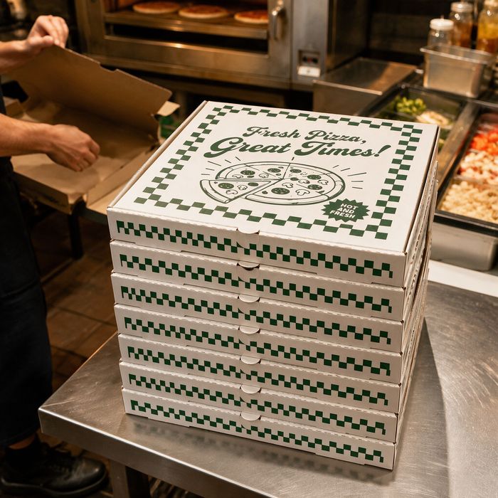 A high stack of commercial pizza carton boxes placed on a stainless steel counter inside a professional pizzeria kitchen environment.
