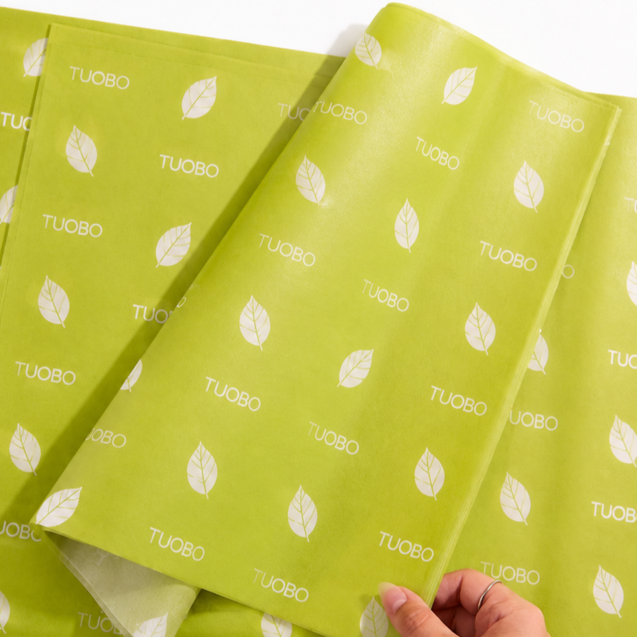 A hand lifting a sheet of custom lime green wrapping tissue paper featuring white leaf illustrations and a white TUOBO logo pattern.