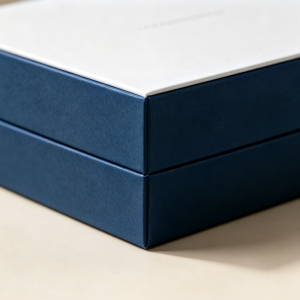 Close-up detail of a premium rigid drawer box, showing tight corner stitching and seamless fit between the white top and navy blue sides, on a neutral beige background.