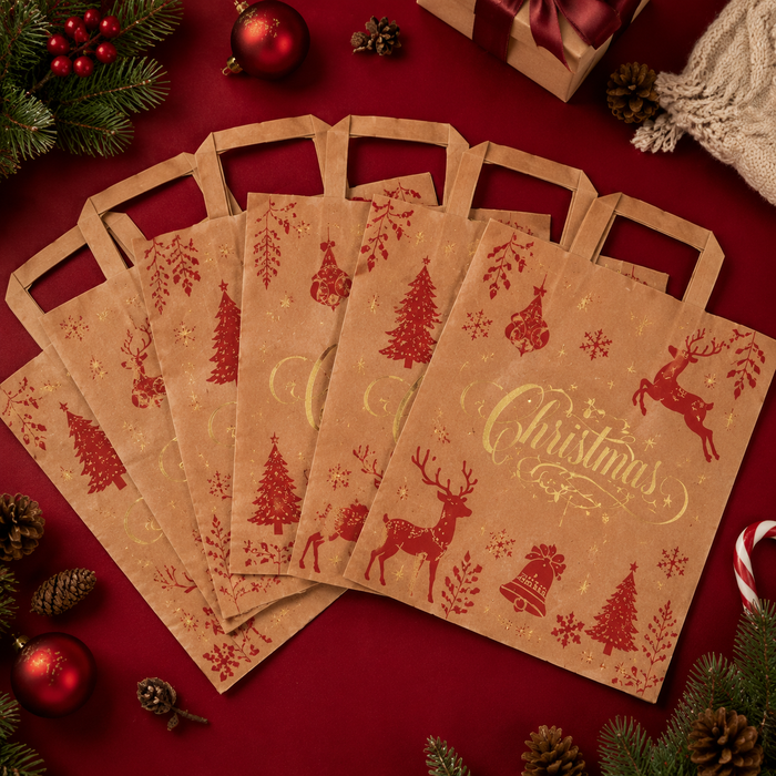 Top-down view of multiple brown kraft paper Christmas gift bags with red and gold festive designs, neatly arranged flat on a red surface with pinecones and holiday accents.