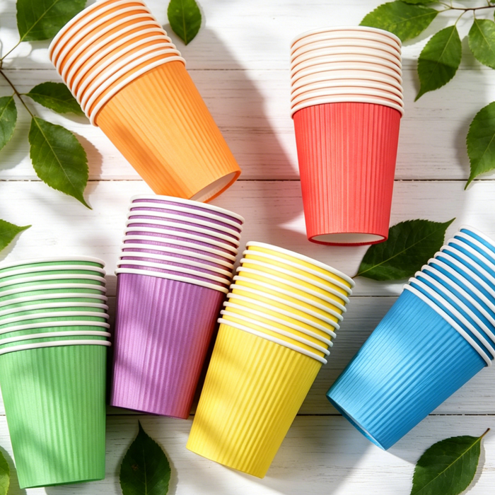 Top view stacked multi-color ripple wall corrugated paper cups, pastel orange red purple yellow green blue insulated disposable party cups on white wood table with fresh green leaves, eco food grade single use hot drink paper cups for birthday wedding holiday wholesale supply