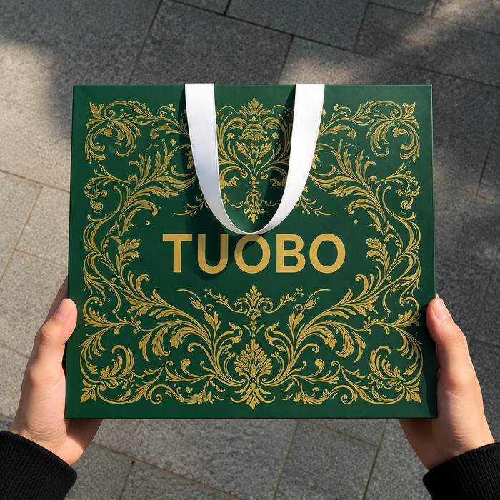Real hands holding dark green embossed gold foil paper bag, outdoor grey pavement daylight shot, reusable custom logo carry bag for clothing & cosmetic boutique wholesale